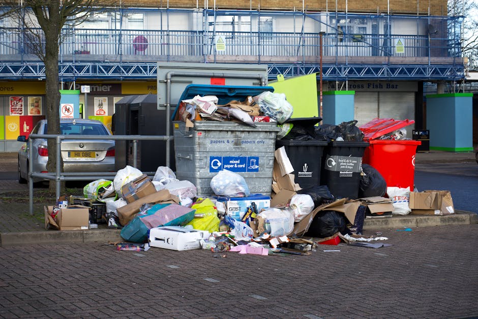 A collection of overflowing rubbish bins and waste materials positioned on a paved urban sidewalk near a parking area. The scene includes a large grey mixed waste skip filled with assorted items such 