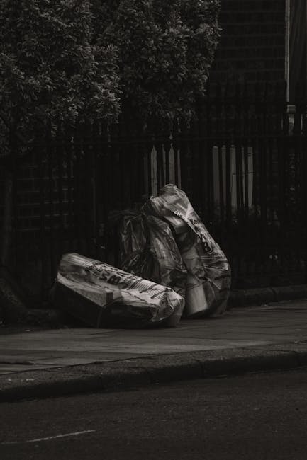 A collection of overflowing rubbish bins and waste materials positioned on a paved urban sidewalk near a parking area. The scene includes a large grey mixed waste skip filled with assorted items such 