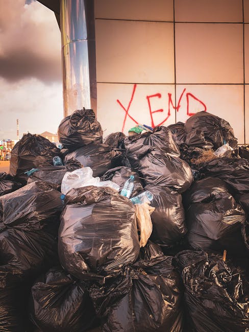 A low-angle view of an outdoor rubbish collection point showing a mix of discarded waste materials. In the foreground, a worn, beige car tire leans against a pile of black and white plastic garbage ba