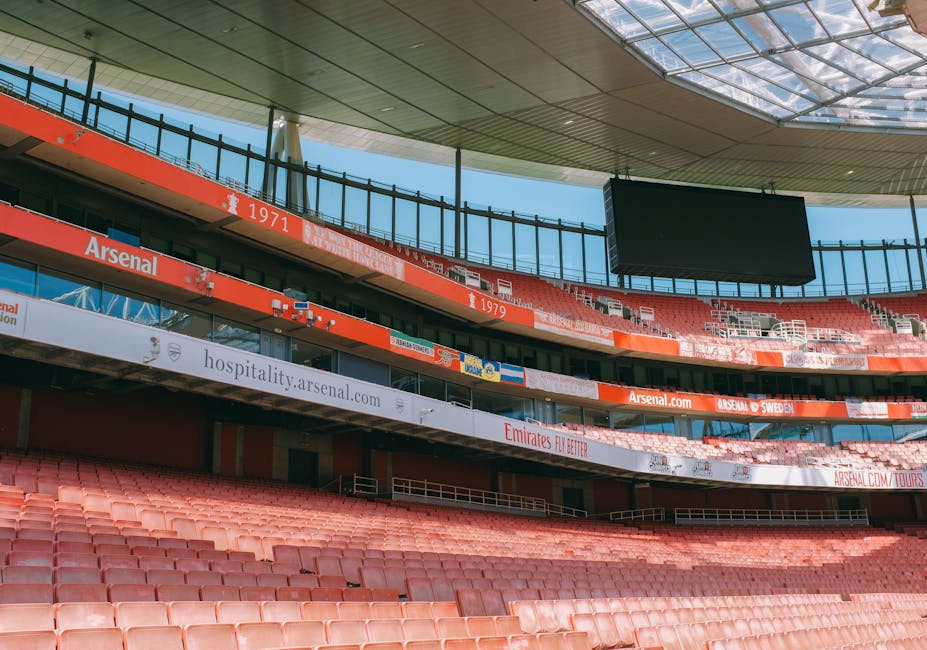 An empty section of stadium seating with rows of weathered red plastic chairs, some showing signs of dirt and wear, arranged to face the field area. The seats are positioned on a sloped concrete tier,